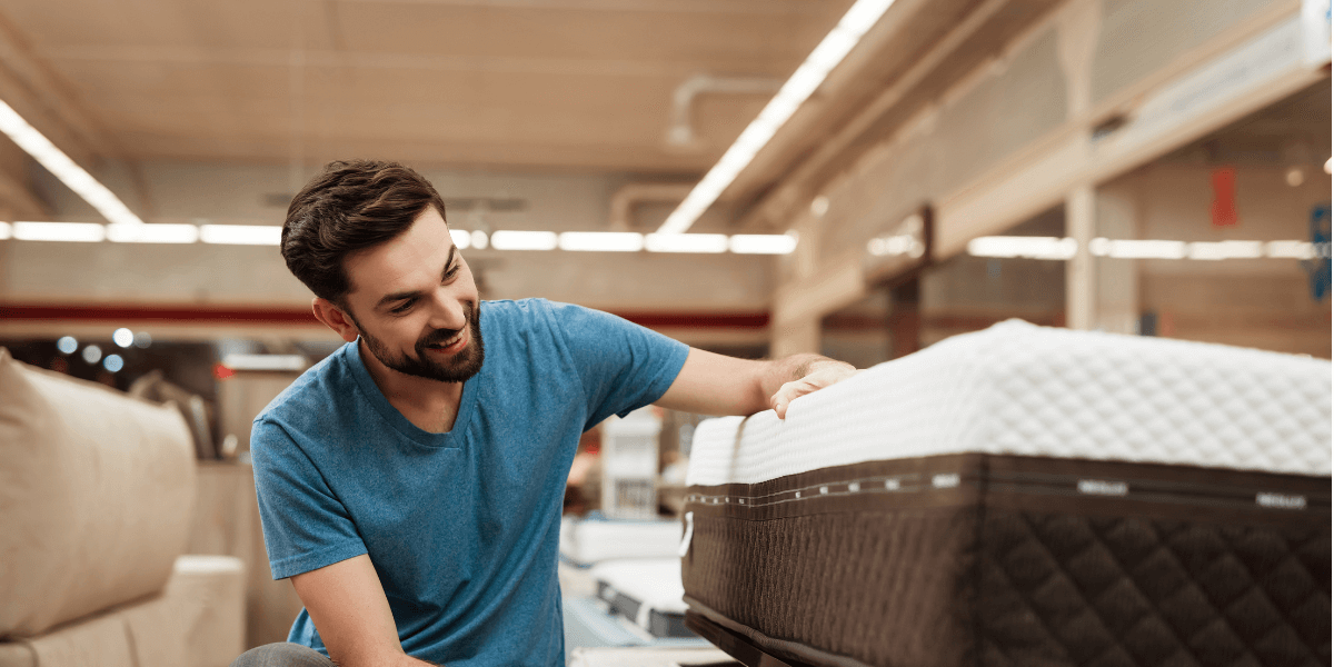 A man stands in a store, inspecting a mattress as he evaluates his choices for bedding.